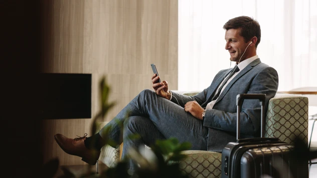 Forretningsreisende i dress sitter avslappet i lounge med koffert og mobil – firmareiser organisert av et reisebyrå for bedrifter. Forretningsreisende i dress sitter avslappet i lounge med koffert og mobil – firmareiser organisert av et reisebyrå for bedrifter.